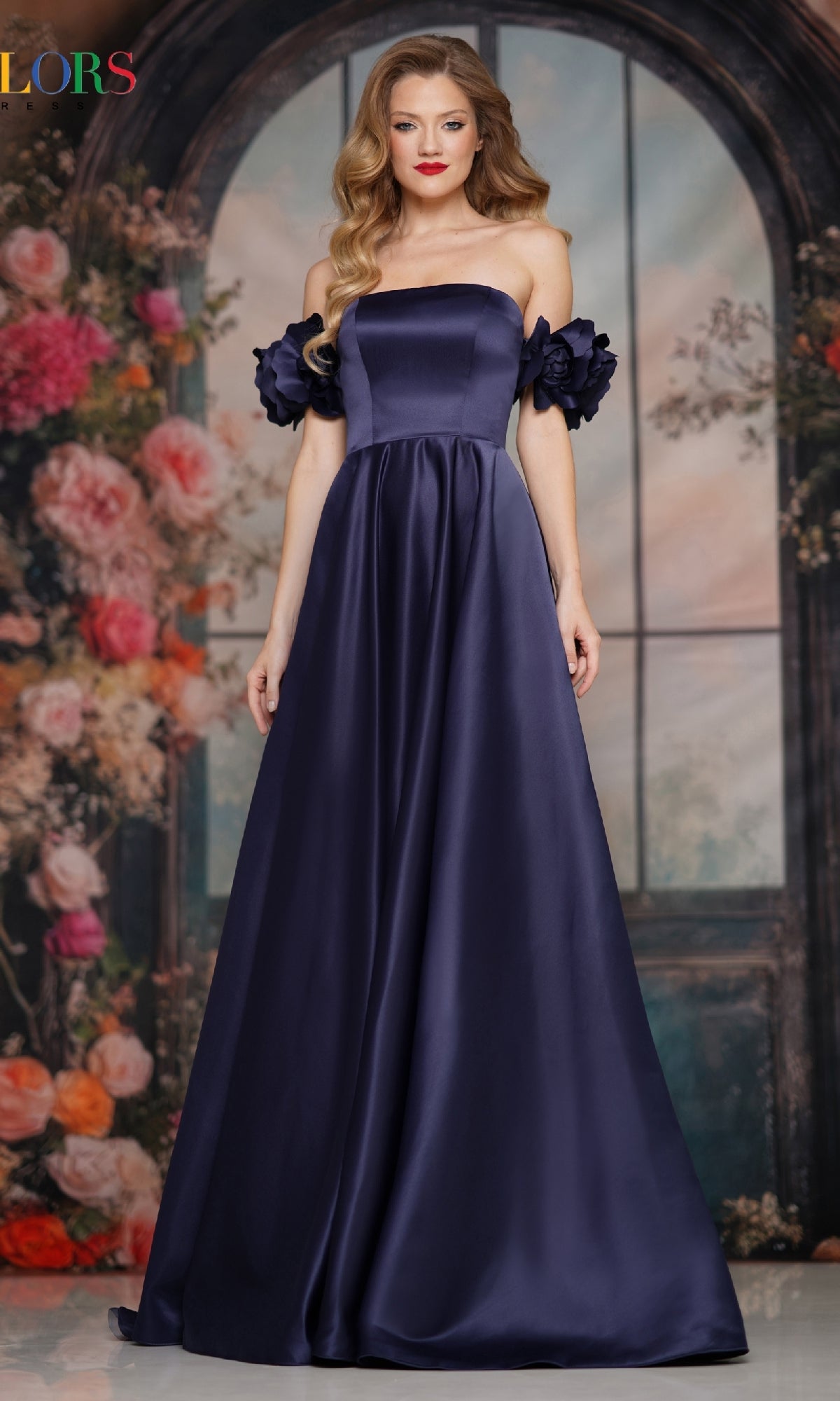 A woman wearing an off-shoulder satin prom gown by Colors Dress, stands in front of a backdrop with colorful flowers and an arched window.